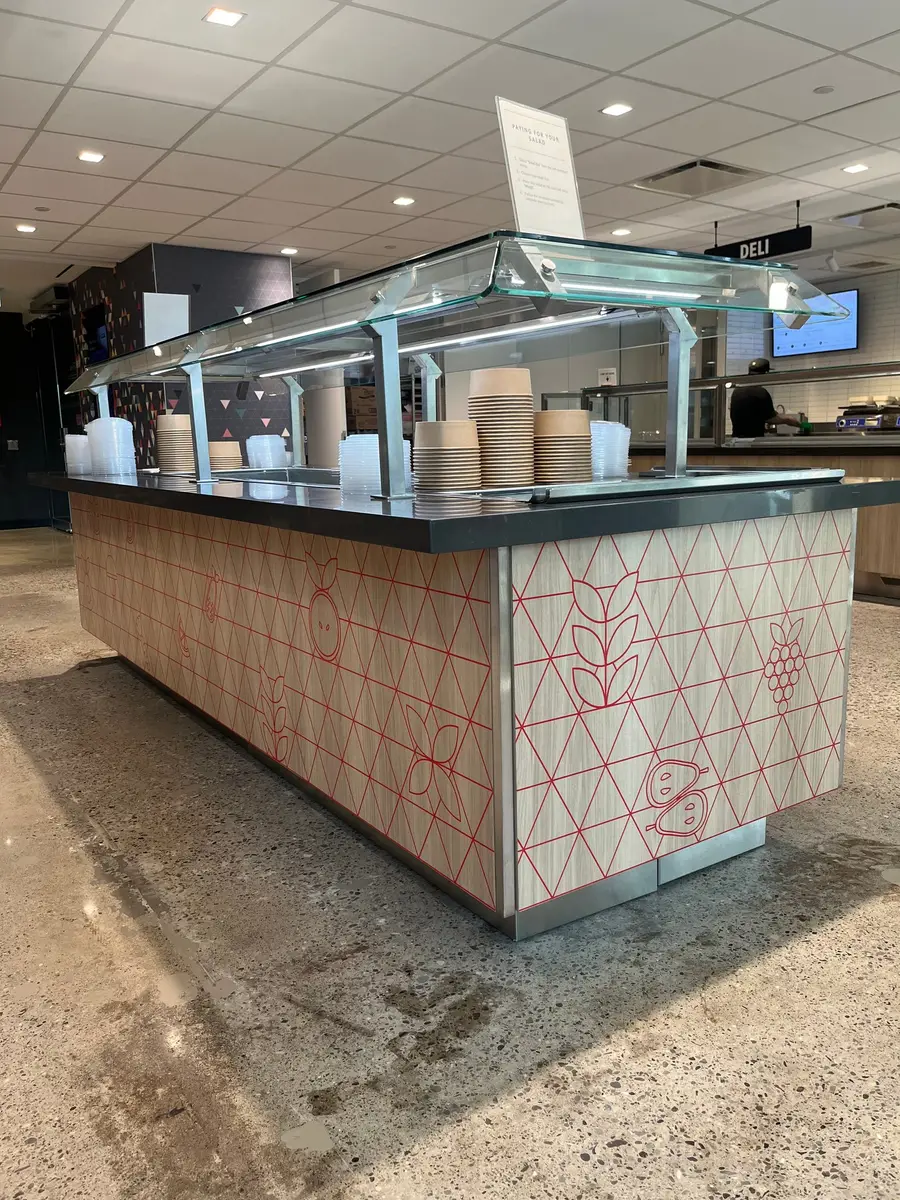 A salad bar station with a glass sneeze guard features a custom graphic wrap on the wood panels depicting red fruit and vegetable line art.