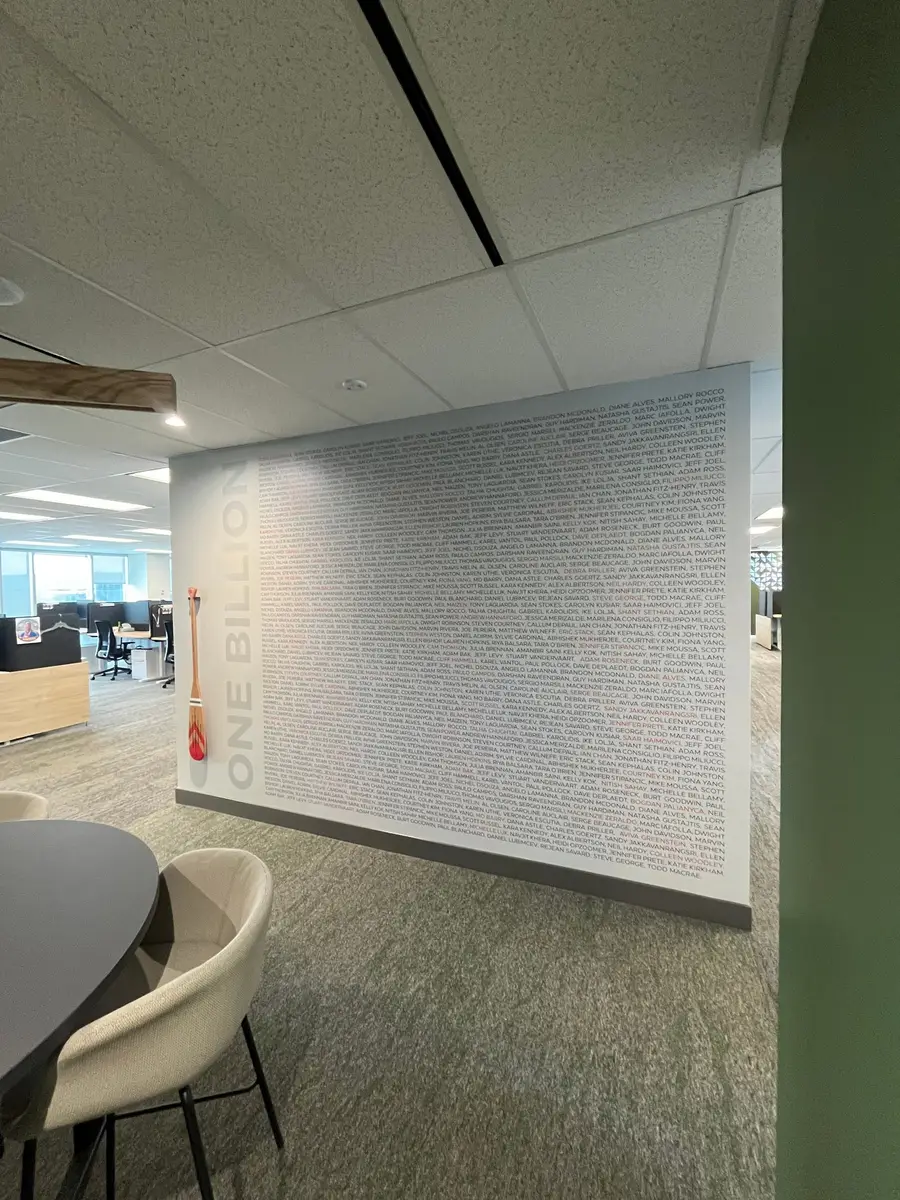 A wide view of an office wall displays a One Billion milestone mural covered with hundreds of employee names and decorative paddles.