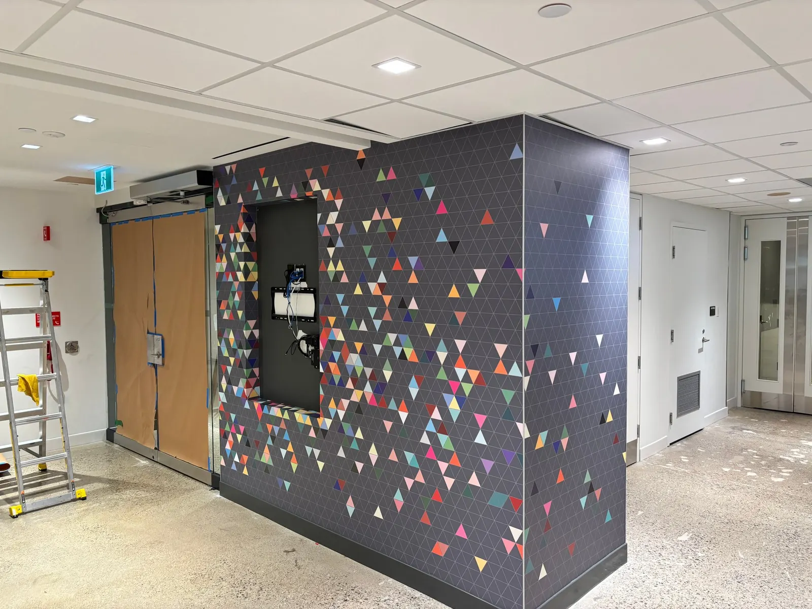 A wide-angle view shows The Hub feature wall with a colorful scattered triangle pattern wallcovering wrapping around a structural column during finishing stages.
