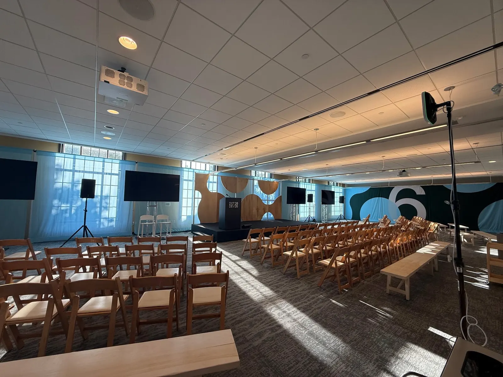 A bright conference session room for DotDev2026 is set with wooden folding chairs, a stage, and geometric branded backdrop panels.