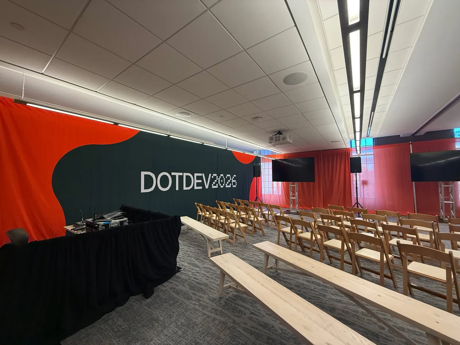 A breakout session room features a red and green DotDev2026 branded backdrop, wooden chairs, and display screens.