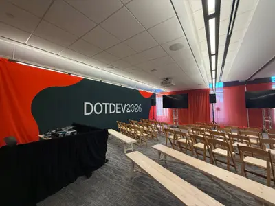 A breakout session room features a red and green DotDev2026 branded backdrop, wooden chairs, and display screens.