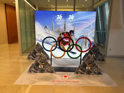 A large LED screen showing Milano Cortina 2026 Team Canada imagery alongside an Olympic rings prop and mountain cutouts in a corporate lobby.