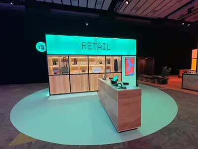 A Shopify Retail trade show booth features teal illuminated signage, wooden shelving displaying merchandise, and a checkout counter.