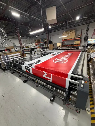 A large-format banner featuring Markham and Brannn branding is being printed on an industrial wide-format printer inside a print shop.