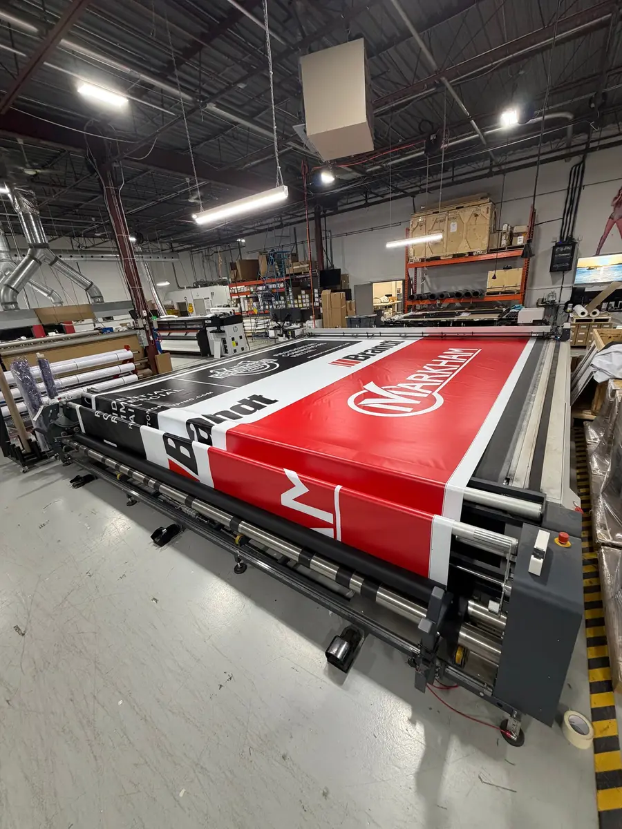 A large-format banner featuring Markham and Brannn branding is being printed on an industrial wide-format printer inside a print shop.