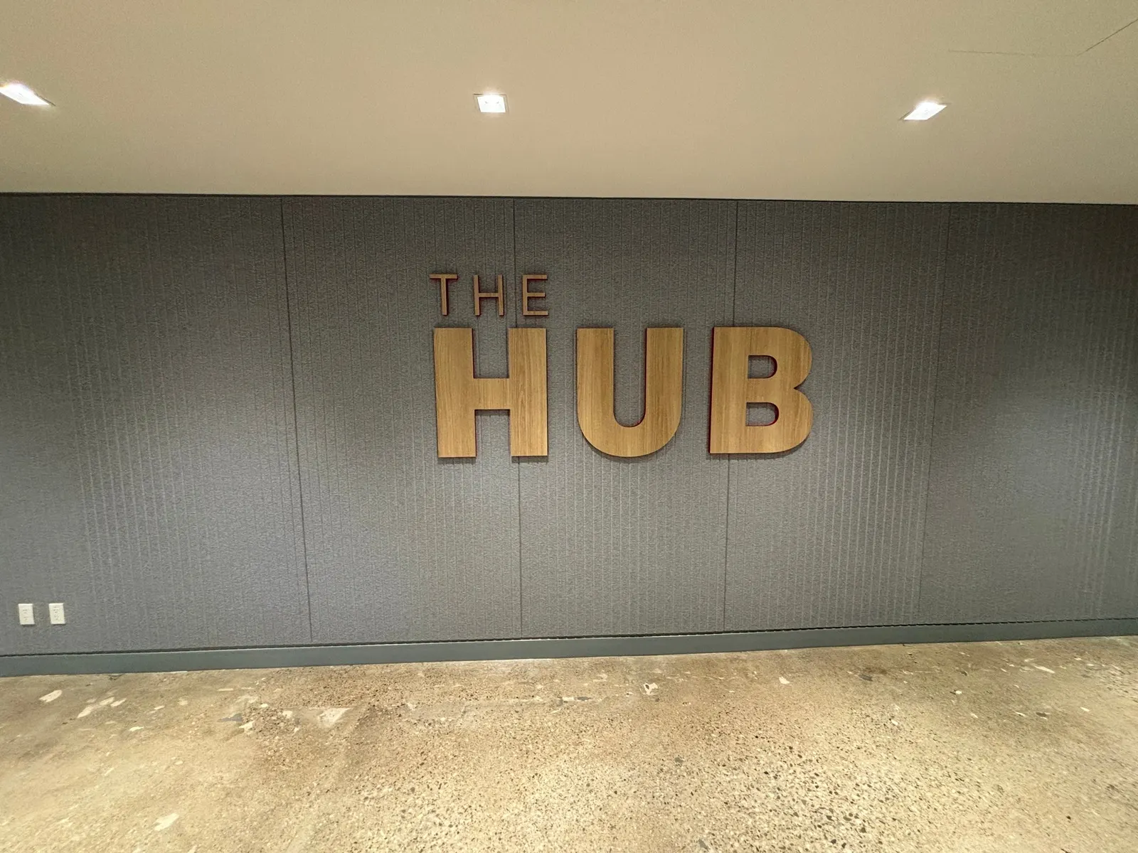 A finished feature wall shows raised wood-grain dimensional letters spelling THE HUB mounted on a dark textured fabric wallcovering.