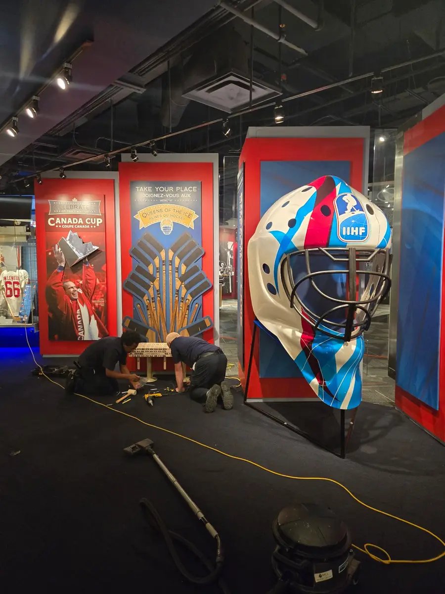 Workers are assembling a hockey stick photo op prop next to an oversized IIHF goalie mask during exhibit installation.