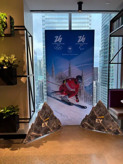 A Milano Cortina 2026 Team Canada vertical banner with mountain props displayed in a high-rise office lounge with a city skyline visible through the windows.