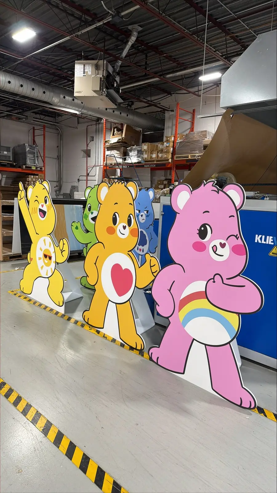 A row of large Care Bears character cardboard cutout standees lined up inside a warehouse fabrication facility.